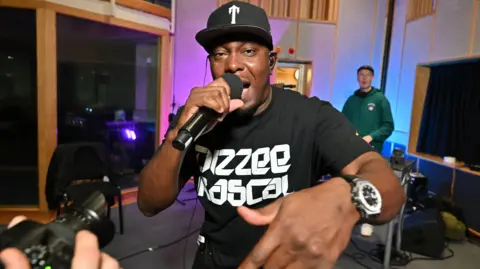 Dizzee Rascal looks at the camera and is wearing a t-shirt with the words Dizzee Rascal on it. He is holding a microphone in one hand and wearing a black baseball cap.