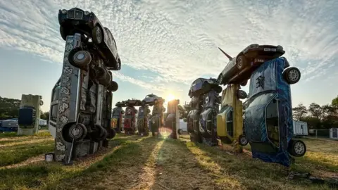 PA Media The image shows Mr Rush's work "Carhenge", which is a replica of Stonehenge made using vintage cars. With the sunrise in the background, 11 cars can be seen wedged vertically in the ground of a field, with others lying on top of them.