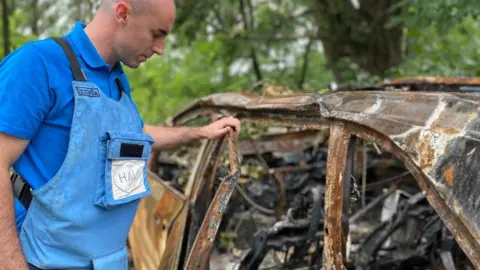 The HALO Trust Kieran standing next to the shell of a burnt out car whilst wearing a safety vest.