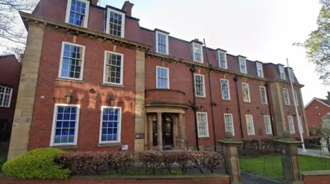 A red-brick building with a white flagpole outside and bushes and a wall