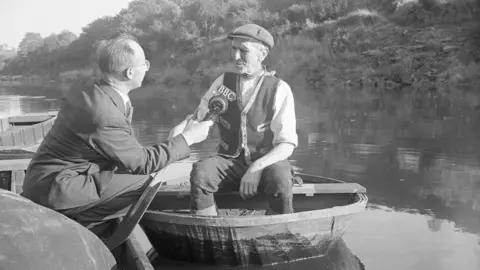 An old black and white photograph of a man wearing a flat cap inside a wooden coracle sitting on the river severn. He is being interviewed by a man wearing a suit holding a BBC microphone
