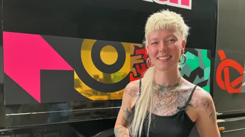 Family Storm's sister is smiling to camera and wearing a black vest top. She has long blond hair at the back which is cut short into a thick fringe at the front and sides. She has tattoos covering her chest and some on her arms and has four chains around her neck and is also wearing big hoop earrings. She is standing in front of a big vehicle with various pink, gold, red, brown, green, black and grey symbols on.