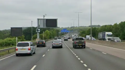 Google A motorway carriage with cars queueing on it. There is a blue sign above the road.