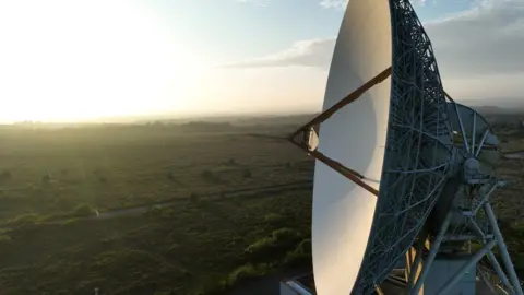 Goonhilly Earth Station A large satellite tracking antenna in a pale colour with a background of green fields and a setting sun.