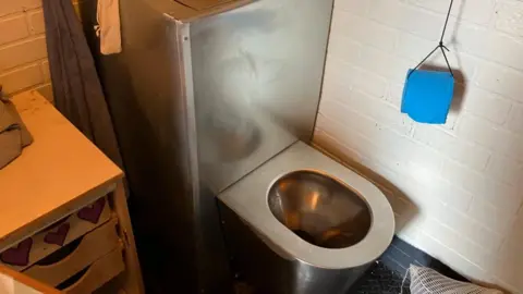BBC/Nicola Rees A metal prison toilet is pictured beside a basic set of drawers. The walls are brick, painted white and there is a blue toilet roll hanging on a piece of wire from the ceiling. There is a towel hanging on the wall next to the toilet.