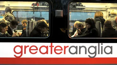 A close-up photo of the outside of a Greater Anglia train. It shows passengers looking at their phones, taken in Dec 2023. 