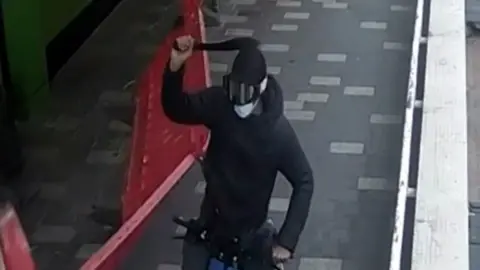 Bedfordshire Police A CCTV still of one of the brothers holding a large knife above their head. He has his face covered by goggles and a blue mask. He is on a bike and wearing all black. Next to him is a plastic board that has been knocked over.