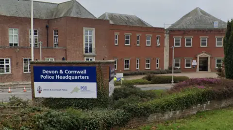 The police headquarters has a sign in front of it reading Devon and Cornwall Police Headquarters, Devon and Cornwall Police and PCC. The buildings are two storey made of red brick. There is planting in the foreground.