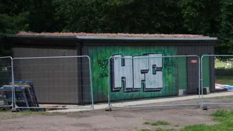 One wall of the toilet block has been spray painted green and there are three large letters on the side "HFC". There is a workmen's temporary fence around the block.