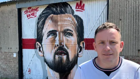 John Devine/BBC Street artist Nathan Murdoch looking at the camera. His mural of Harry Kane is behind him on a building.