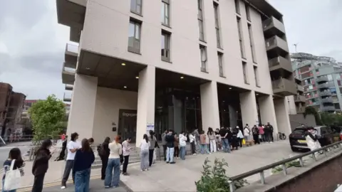 A long queue of people underneath a big cream block of flats.