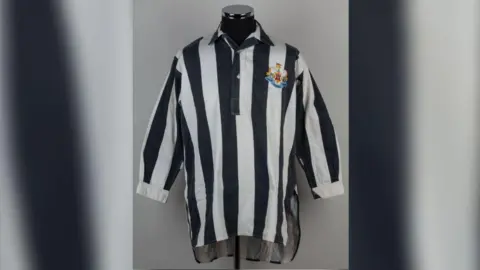 The 1952 Black and white shirt Newcastle shirt displayed on mannequin. It has a Newcastle logo on the right hand side and long sleeves.