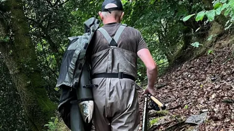 Natural Resources Wales Samuel Stephen, wearing dungarees, is seen from behind with a whole salmon protruding from the sleeve of a coat slung over his shoulder