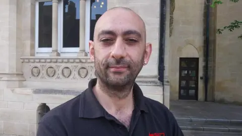 Nadia Lincoln/LDRS Close-up of a man with a stubble and pierced ear standing outdoors in front of a light stone building with decorative columns.