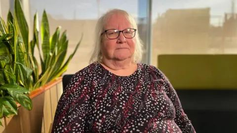 A woman with white hair just above her shoulders who is wearing black rectangular glasses. She is sat in a chair looking into the camera with a serious expression. She is wearing a spotty-striped dark red long-sleeved top. Behind her is a snake plant and a long green and black chair.