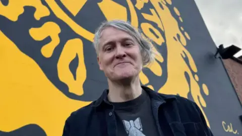 BBC/John Bray A man with greying hair swept across his forehead stands in front of a yellow and black mural on the side of a house. He is wearing a black suede jacket and black t-shirt with white graphic.