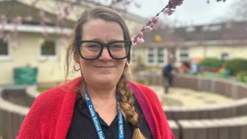 Aimee Dexter/BBC Clare Laidler is off centre to the left and is smiling. She has blonde hair in a plait and is wearing black framed glasses, a black top and a pink and red cardigan. She is standing in a small garden which is situated in between a building.