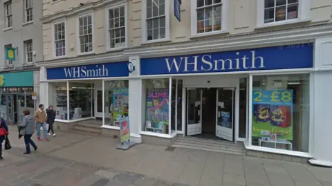 Google WH Smith photographed outside from the pavement, with a number of people walking by the store. 