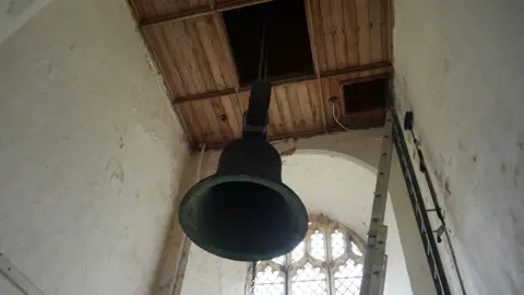 Shaun Whitmore/BBC A dark-coloured church bell suspended above a church ceiling, with a hole in the ceiling directly above it 