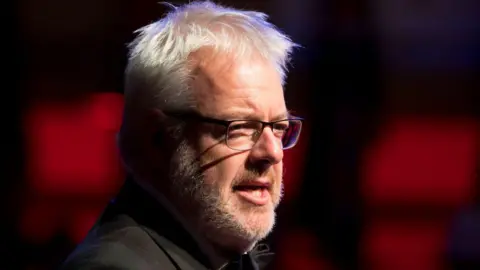 carwyn looking to the right almost in profile in front of a blurry red background. he had a white beard and white shot cropped hair