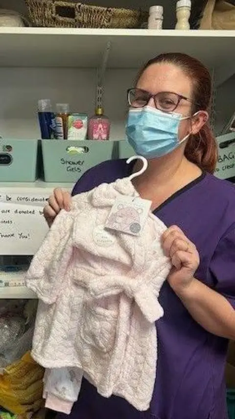 Bradford Teaching Hospitals NHS Foundation Trust A nurse wearing a blue face mask holds up a baby outfit while standing in a cupboard filled with boxes of toiletries.