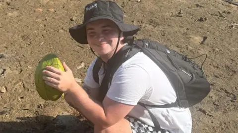 Ashton Taylor Ashton Taylor, with a black sun hat on, wearing a black rucksack is crouched down holding a large piece of fruit