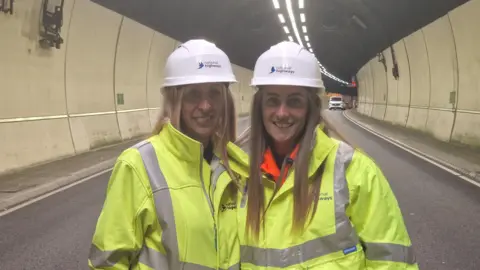 A mother and daughter both with long blonde hair stood in the centre of a dual carriageway in a tunnel. The road is clear of vehicles apart from a white van in the background on the right. The women are wearing yellow high-vis jackets and white hard hats while smiling at the camera