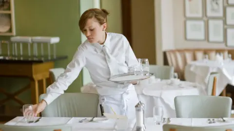 Getty Images A waitress dressed in a white shirt, white tie and white apron sets out water glasses in restaurant
