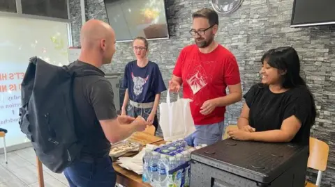 Aishah Ahmed Aishah with volunteers at a table in her food bank. There are water bottles and food on the table and one of the volunteers is handing a man a plastic bag.