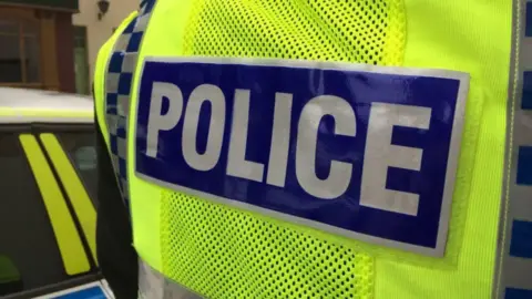 The back of a police officer wearing a bright yellow hi-vis jacket with "police" in capitals in silver and blue. The side of a police car is next to them.