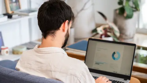 Getty Images Young man with dark hair and beard sitting on a blue couch in beige shirt with his back to the camera, looking at a laptop screen showing a financial report with graphics