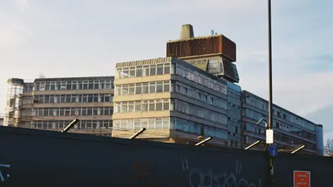 Sovereign House in Norwich is visible behind security hoardings, with its spiral glass staircase, concrete and glass offices and unique tower structure on top visible