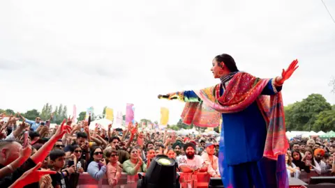 Sandwell & Birmingham Mela A woman in a sari stands on stage and holds a microphone towards the audience.