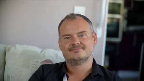 Jamie Niblock/BBC A man is sitting on a sofa and is wearing a black shirt. He is looking directly at the camera and smiling. He has short dark grey hair and a beard.