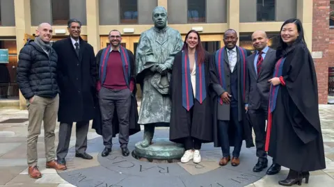 A posed group shot featuring students who took part in the specialist kidney training, together with consultants who created the court. They are five men and two women lining up either side of a metal statue outside Newcastle University.
Four of them, two men and two women, wear a graduation gown with a blue and red and blue stole. The other three men are dressed in formal wear.