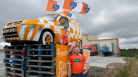 A man and a boy in front a van that is up on pallets. The van is painted orange and white and has orange and white flags on it. 