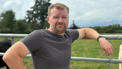 John Story leans on a metal fence, wearing a grey t-shirt. He stands in front of a field with cattle.