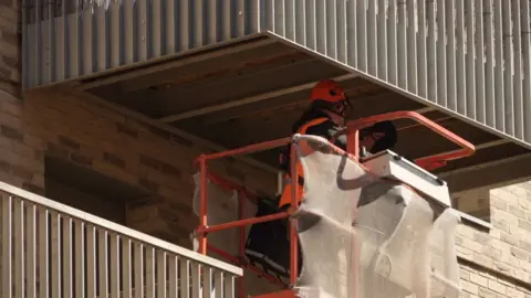 Two engineers in orange helmets and black uniforms inspect the underside of a balcony from a cherry picker