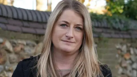 Shaun Whitmore/BBC Det Insp Alix Wright, with long blonde hair and wearing a black coat and brown top, stands in front of a flint wall 