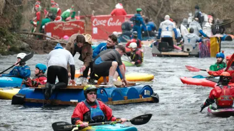 Sam Hepburn Kayakers can be seen on a river in between custom-made rafts with multiple people on top of them. The rafts are made from blue barrels bound together by wood.