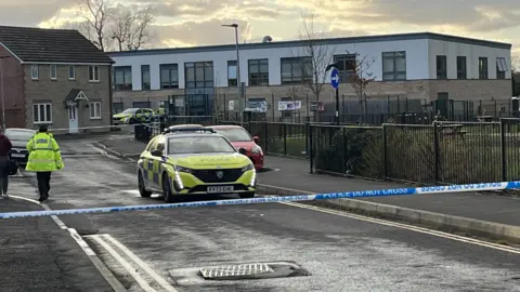 BBC In the foreground, there is blue and white police tape that says "do not cross". In the background, a school, a police car and a police officer with his back to the camera in their hi-vis yellow uniform 