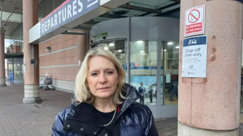 Deputy Elaine Millar, a woman with blonde hair, wearing a navy blue puffa jacket over a navy blue jumper. She has a golden necklace around her neck. A sign saying 'Departures' is visible above her head. Behind her are revolving doors - the entrance to Jersey Airport. To the right of the image is a sign on a pillar, which says 'Drop off zone only'. Another sign above it says 'No smoking in this area'.