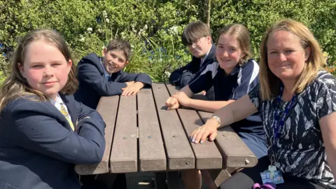 A number of pupils and one member of staff are sat around a wooden bench, all looking at the camera. One one side of the bench, a boy and a girl are sat. They are both wearing navy blue school blazers. On the other side of the bench a member of staff is wearing a black and white patterned top with two pupils sat next to her. One is a girl wearing a navy and white PE top and the other is a boy wearing black framed glasses and a navy blazer. 
