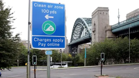 A view of a blue 'Clean air zone' sign in Newcastle city centre. It says 'Charges apply pay online'. Traffic lights on green can be seen behind the sign and a large metal bridge in the background.