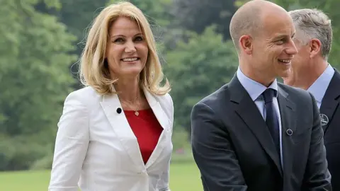 Getty Images Helle Thorning-Schmidt and Stephen Kinnock Two walk across a lawn at an official event. She is wearing a white suit with a red top, while he is dressed in a dark suit and tie. A third suited person is partially visible in the background. Trees and open green space can be seen behind them.