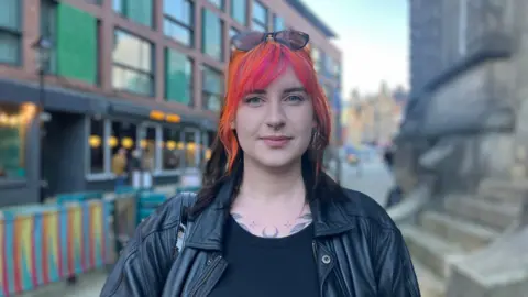 Elizabeth Baines / BBC A woman with orange hair stands in front of the Corn Exchange in Leeds. She wears a black top and black leather jacket.