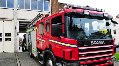 A fire engine which is mainly red with a silver back outside a fire station. 