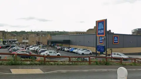 A google streetview image showing an Aldi supermarket. It looks like a small dark-grey warehouse. The car park outside is full.