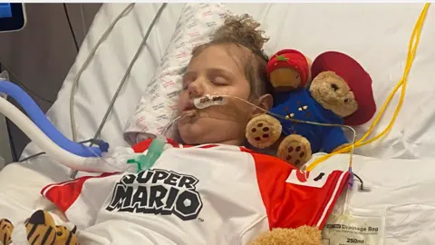 Little boy with tubes in his nose and arms on hospital bed and eyes closed surrounded by teddy bears