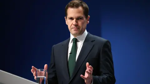 EPA Image of Jenrick. He is at a lectern giving a speech in a green tie and navy suit.